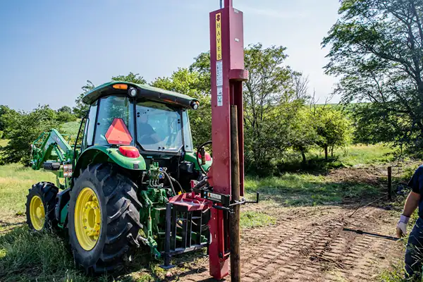 Tractor-mounted post driver installing fence posts on a farm pasture.